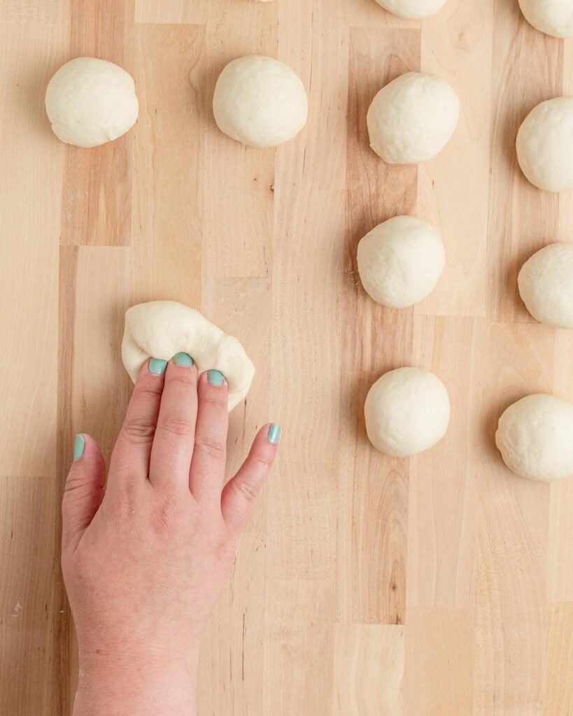 A hand with aqua-painted nails shapes a ball of dough on a wooden surface. Eleven other dough balls are arranged nearby, ready for baking or further preparation.