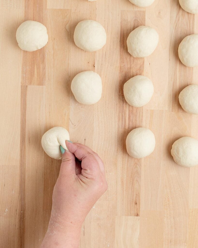 A hand pinches a ball of dough on a wooden surface. Several other dough balls are arranged nearby, ready for baking or further preparation.