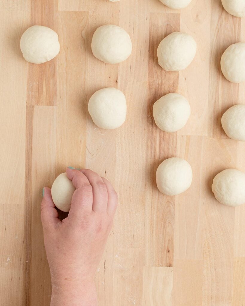 A hand is holding a small ball of dough on a wooden surface, with several other dough balls arranged nearby.