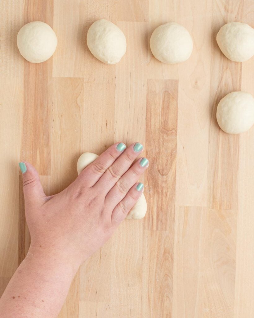 A hand with teal nail polish presses down on a ball of dough on a wooden surface. Five other dough balls are arranged nearby.
