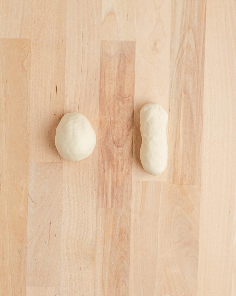 Two pieces of dough rest on a light wooden surface. The dough on the left is round, while the dough on the right is oval-shaped. The wood shows natural grain patterns in varying light brown tones.
