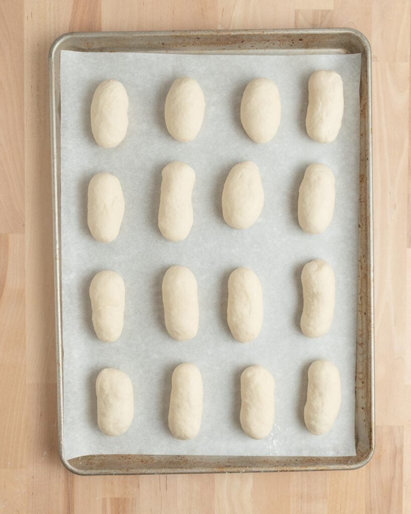 A baking sheet lined with parchment paper holds fifteen evenly spaced, oval-shaped pieces of dough before baking, arranged in a 3 by 5 grid on a wooden surface.