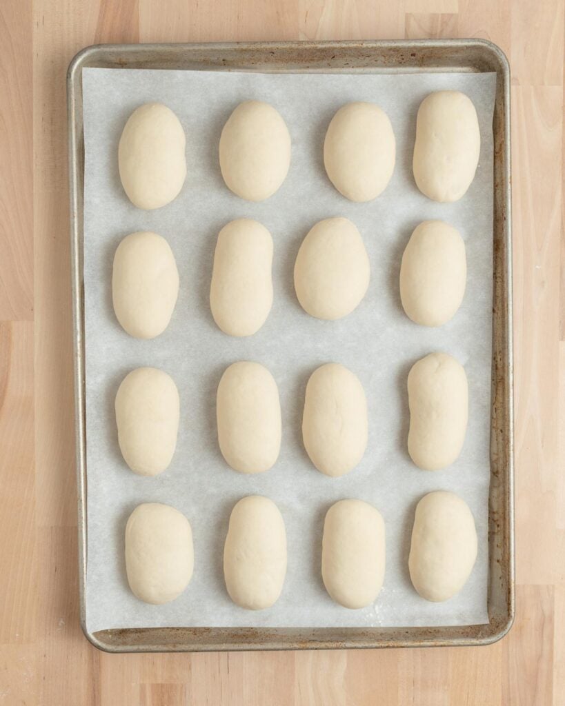 A baking sheet lined with parchment paper holds sixteen evenly spaced, oval-shaped balls of raw dough, arranged in four rows of four, on a light wooden surface.
