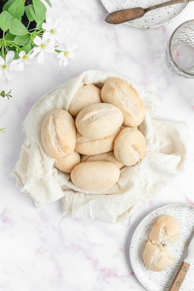 A basket lined with a white cloth holds several oval bread rolls on a marble surface. Nearby are a glass of water, a butter knife, a plate with a roll, and white flowers in a pot.