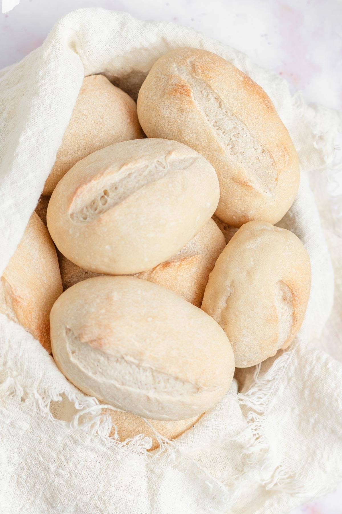 Seven round, light brown bread rolls with a split top sit in a white cloth-lined basket on a white surface.
