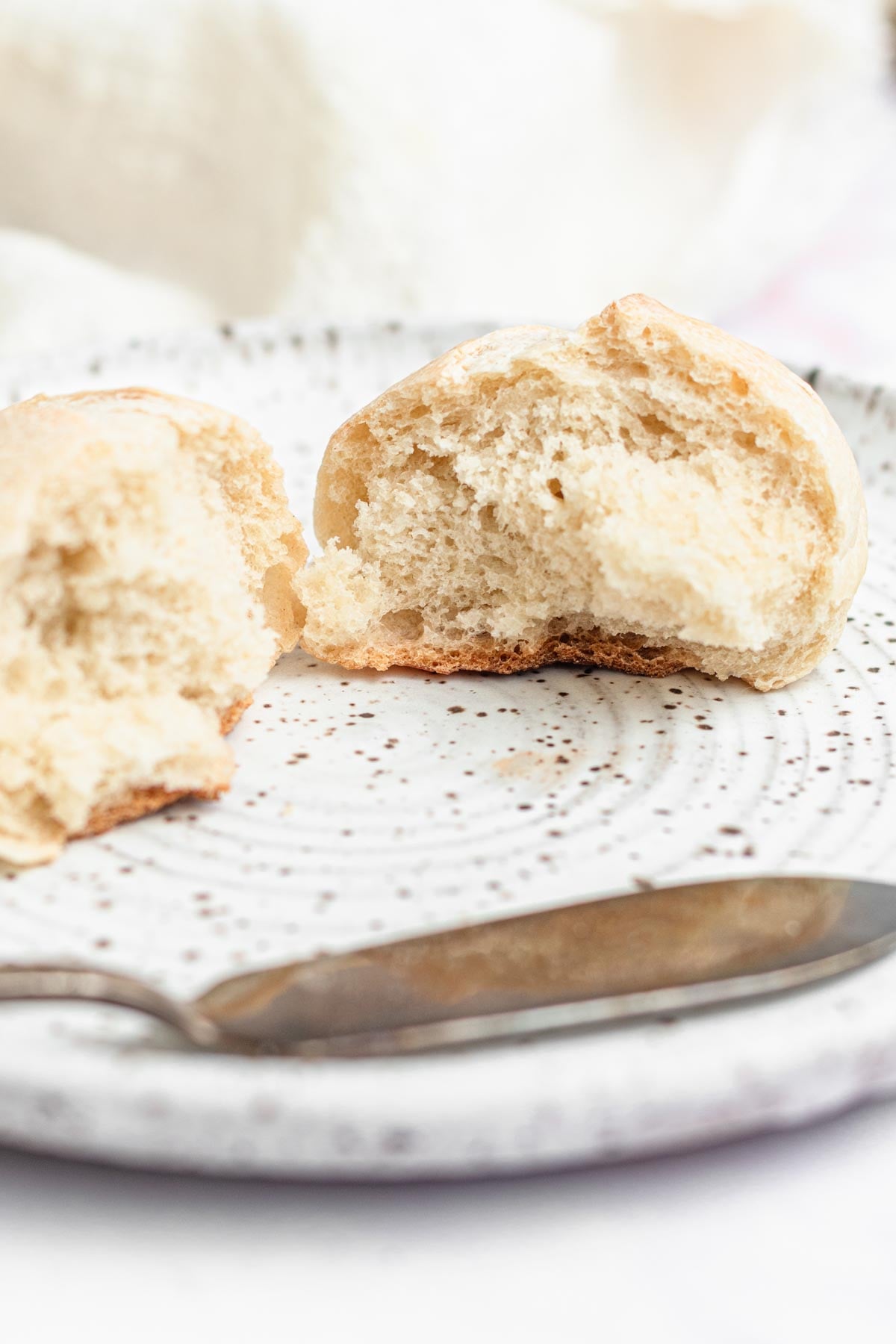 A round bread roll is split in half on a white speckled plate. A butter knife rests nearby. The rolls inside texture is visible, and the background is softly blurred.