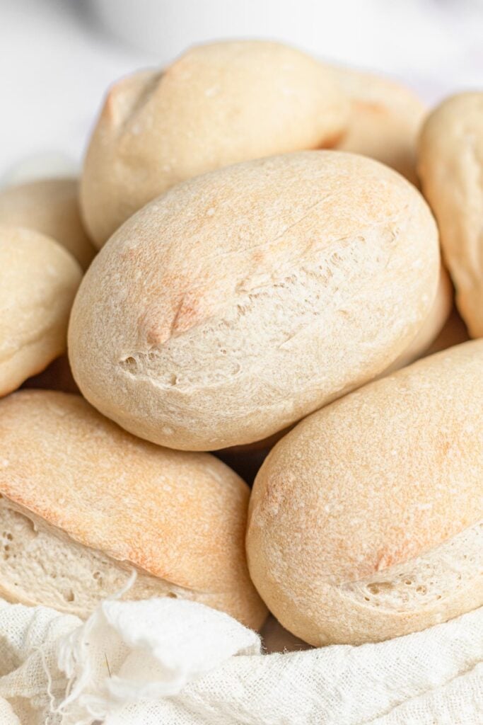 A close-up of several oval-shaped, lightly browned bread rolls stacked together in a basket lined with a white cloth.