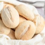 A close-up of several oval-shaped french bread rolls with light brown crusts and slits on top, arranged in a cloth-lined basket.