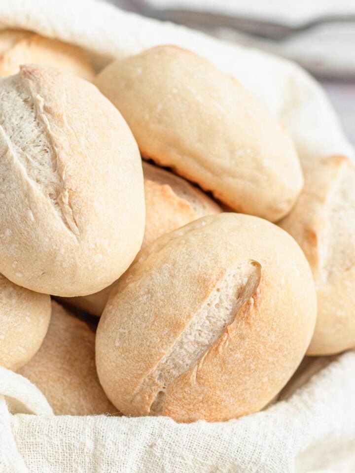 A close-up of several oval-shaped french bread rolls with light brown crusts and slits on top, arranged in a cloth-lined basket.